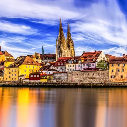 Historische Regensburger Skyline in Bayern mit Domtürmen und bunten Gebäuden, die sich in der Donau unter dramatischem Himmel spiegeln.