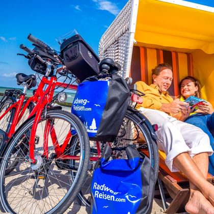 Zwei Radfahrer entspannen in einem gelb-orangen Strandkorb am Ostseestrand in Wustrow, mit roten Fahrrädern daneben unter blauem Himmel.