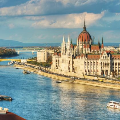 Budapester Parlamentsgebäude mit roter Kuppel am Donauufer. Blauer Fluss mit Booten, Brücke und fernen Bergen unter bewölktem Himmel.