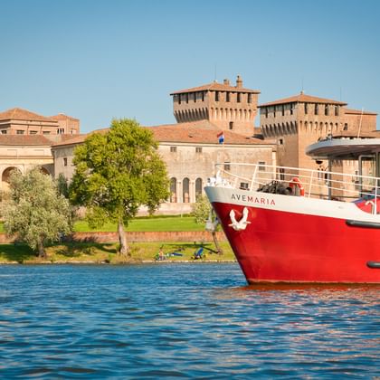 Red and white ship MS Ave Maria on blue water with historic Mantua fortress and towers in the background under clear sky.
