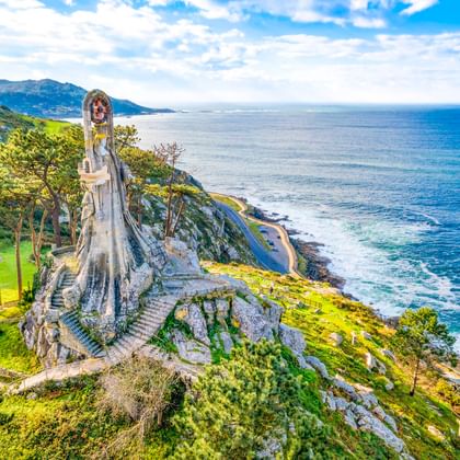Virxe da Roca Steinmonument auf Küstenklippe in Baiona mit Blick auf den Atlantik, grüne Hänge und entfernte Berge.