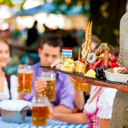 Kellner in traditioneller bayerischer Tracht hält Tablett mit Käse, Brezeln und Snacks. Gäste in Dirndl und Lederhosen mit Bier im Hintergrund.