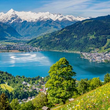 Türkisfarbener Alpensee umgeben von grünen Wiesen mit gelben Wildblumen, bewaldeten Hügeln und schneebedeckten Berggipfeln unter blauem Himmel.