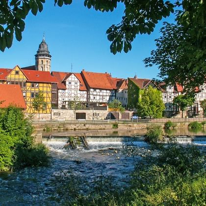 Fachwerkhäuser mit roten Dächern und Kirchturm an der Weser. Ein Wehr erzeugt fließendes Wasser im Vordergrund, eingerahmt von grünen Bäumen.