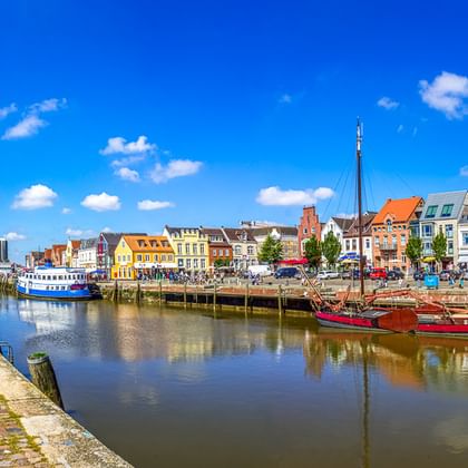 Historischer Hafen von Husum mit bunten Häusern entlang der Uferpromenade. Rote und blaue Boote liegen am Kai unter blauem Himmel.