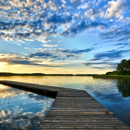 Holzsteg, der sich bei Sonnenuntergang in einen ruhigen masurischen See erstreckt. Der Himmel ist voller dramatischer Wolken, die sich im Wasser spiegeln.