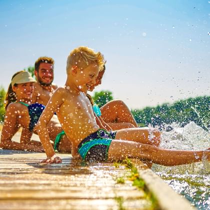 Familie mit zwei Kindern planscht auf einem Holzsteg am Brombachsee, mit klarem blauen Himmel und grünen Bäumen im Hintergrund.
