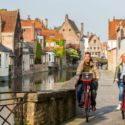Zwei Radfahrer auf roten Rädern überqueren eine Steinbrücke in einer historischen flämischen Stadt mit Kanal, traditionellen Backsteinhäusern und Ziegeldächern.