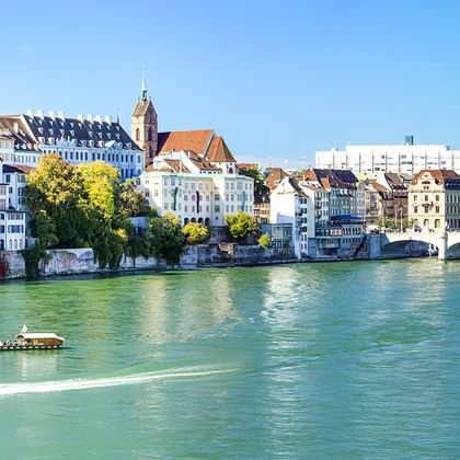 Grüner Rhein mit Booten, historischen Basler Gebäuden, Kirchturm und Steinbrücke unter blauem Himmel. Bäume säumen das Ufer.