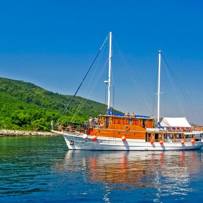 MS Mirabela, ein weißes Segelschiff mit Holzaufbau und zwei Masten, ankert in ruhigem blauen Wasser vor grüner bewaldeter Küste.