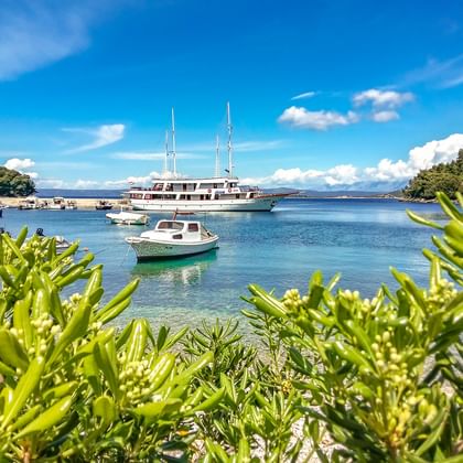 Malerische Bucht in Korcula, Kroatien mit großer Yacht und kleinen Booten im türkisfarbenen Wasser, eingerahmt von grüner Vegetation unter blauem Himmel.