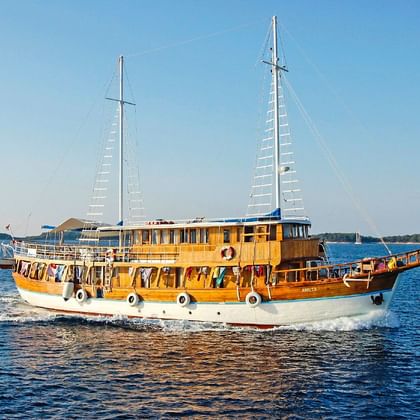 MS Aneta, ein hölzernes Segelschiff mit zwei Masten und weiß-braunem Rumpf, fährt auf ruhigem blauen Wasser mit grünen Inseln im Hintergrund.