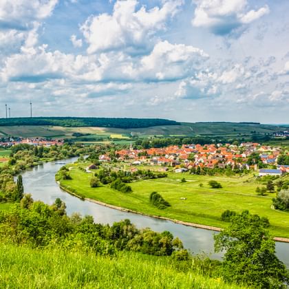 Luftaufnahme eines sich schlängelnden Flusses durch grüne Wiesen mit einem Dorf mit roten Dächern. Hügel und Windräder in der Ferne unter bewölktem Himmel.