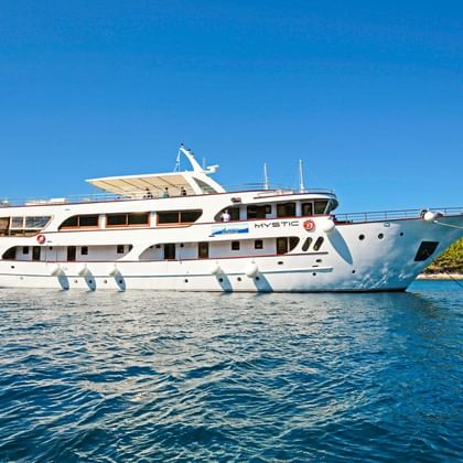 Weiße Motoryacht MY Mystic ankert in ruhigem blauen Wasser mit grüner Küstenlinie im Hintergrund unter klarem Himmel.