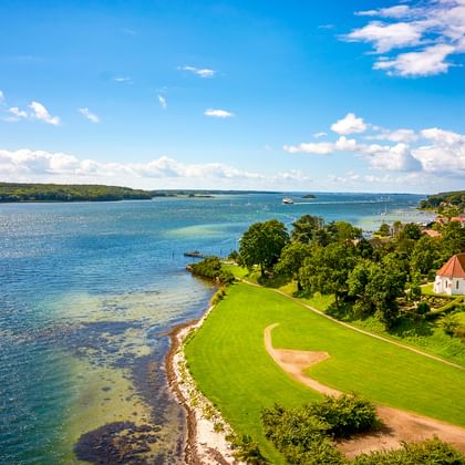 Luftaufnahme der Küste von Svendborg mit weißer Kirche auf grüner Halbinsel, blauen Fjordgewässern und Wohngebiet unter hellem Himmel.