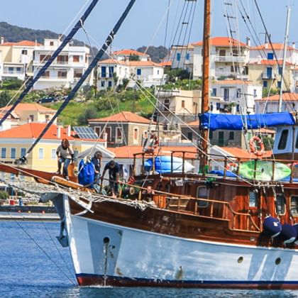 MS Panagiota, ein hölzernes Segelschiff mit weiß-braunem Rumpf, liegt in einem griechischen Hafen vor bunten Gebäuden und Hügeln.