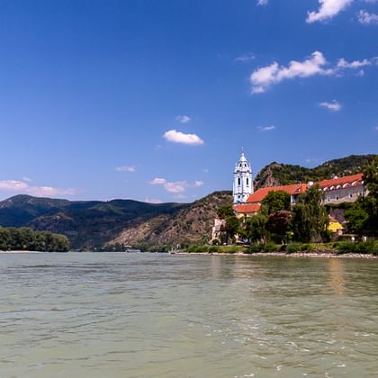 View of the Danube River with Dürnstein Abbey's blue-white tower and castle ruins on a hilltop. Mountains and blue sky in the background.