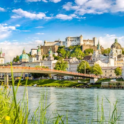 Blick über die Salzach auf Salzburgs Altstadt mit Festung Hohensalzburg auf dem Hügel, historischen Kirchen und Brücke, eingerahmt von grünem Ufer.