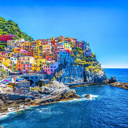 Leuchtend bunte Häuser eines Cinque Terre Dorfes auf felsigen Klippen über dem blauen Mittelmeer unter klarem Himmel.