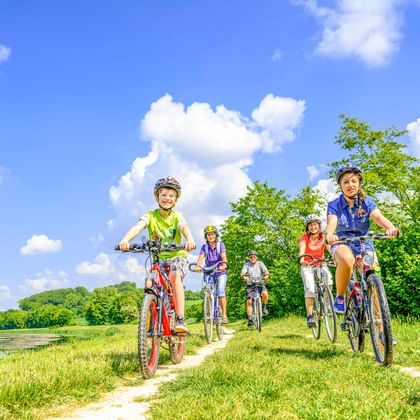 Fünf Radfahrer mit Helmen fahren auf einem Grasweg neben einem Fluss unter blauem Himmel mit weißen Wolken. Grüne Bäume und Hügel im Hintergrund.