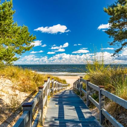 Holzsteg mit Geländer führt durch Sanddünen zum Ostseestrand auf Usedom. Kiefern rahmen den Weg unter blauem Himmel mit Wolken.