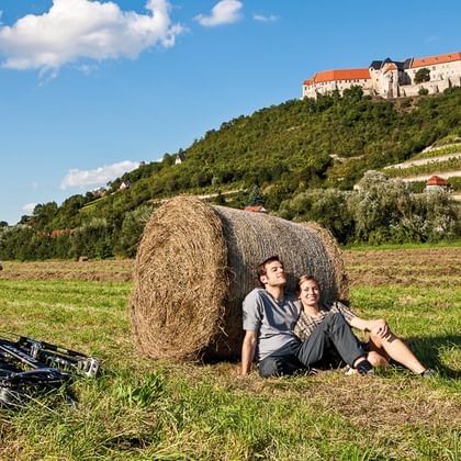 Zwei Radfahrer lehnen an einem Heuballen auf einer Wiese, Fahrräder daneben. Weinberge und eine Burg auf dem Hügel im Hintergrund.