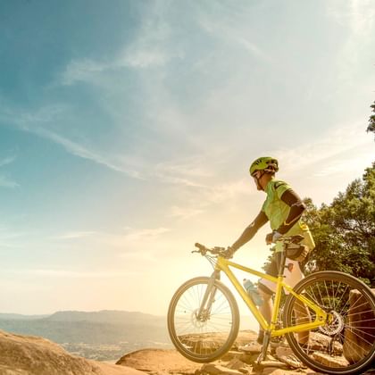 Radfahrer in grünem Trikot und Helm mit gelbem Mountainbike auf steinigem Aussichtspunkt mit Blick auf Tallandschaft mit Bäumen unter blauem Himmel.