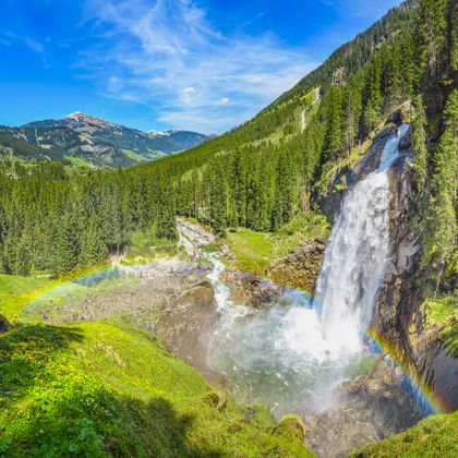 Krimmler Wasserfälle stürzen über Felsklippe mit Regenbogen im Sprühnebel, umgeben von grünen Wiesen und dichten Wäldern in den Alpen.