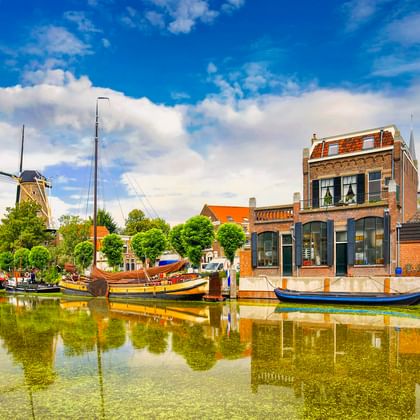 Traditionelle Boote liegen in einem Kanal in Gouda vor historischen Backsteinhäusern und einer Windmühle. Die Szenerie spiegelt sich im ruhigen Wasser.