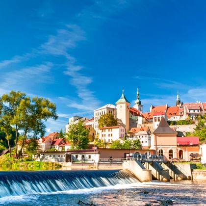 Burg Loket auf einem Hügel über der Eger mit Wehr im Vordergrund. Historische Stadt mit rotgedeckten Gebäuden und Kirchtürmen unter blauem Himmel.