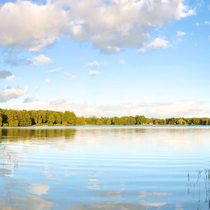 Ruhiger See mit spiegelglatten Reflexionen von Wolken und Himmel. Dichter grüner Wald säumt beide Ufer unter blauem Himmel mit weißen Wolken.
