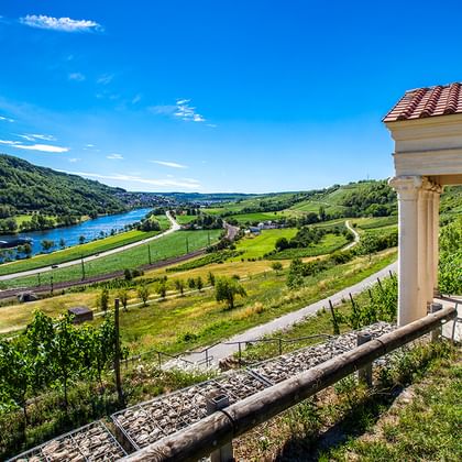 Malerischer Blick auf die Mosel, die sich durch grüne Hügel und Weinberge schlängelt, mit einem Pavillon mit Ziegeldach und weißen Säulen im Vordergrund.