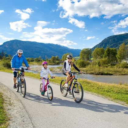 Familie mit drei Personen radelt auf asphaltiertem Weg entlang der Mur mit Bergen im Hintergrund. Erwachsene und Kind tragen Helme an sonnigem Tag.