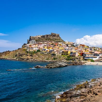 Bunte Hangstadt mit Festungsruinen auf dem Gipfel mit Blick auf eine blaue Mittelmeerbucht mit felsiger Küste in Nordsardinien.
