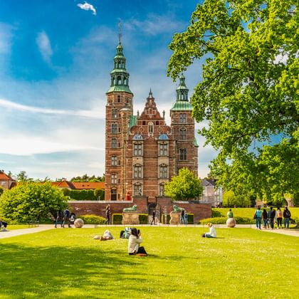 Schloss Rosenborg mit roten Backsteintürmen und grünen Kupferspitzen in Kopenhagen, umgeben von Parkrasen mit Besuchern unter blauem Himmel.