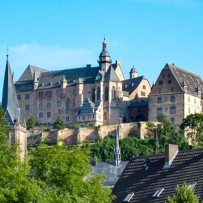 Historic Marburg Castle perched on a hilltop with stone walls and towers, surrounded by green trees. Town rooftops visible below against a clear blue sky.