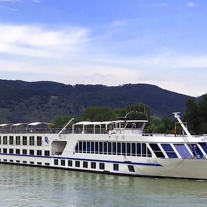 White river cruise ship with blue accents sailing on the Danube. Green hills and mountains visible in the background under a blue sky.