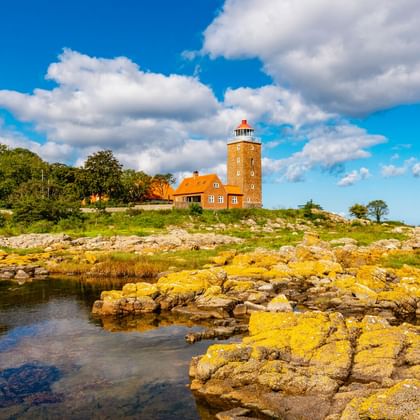 Leuchtturm Svaneke mit rotem Ziegeldach auf felsiger Küste in Bornholm. Gelb bewachsene Felsen im Vordergrund, blauer Himmel mit Wolken.