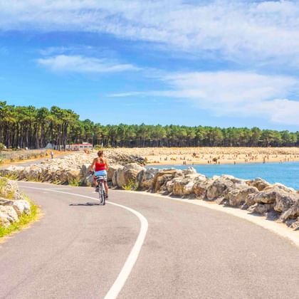 Radfahrerin im roten Shirt auf asphaltiertem Küstenweg bei La Palmyre. Felsige Küste, blaues Wasser, Sandstrand und Pinienwald im Hintergrund.