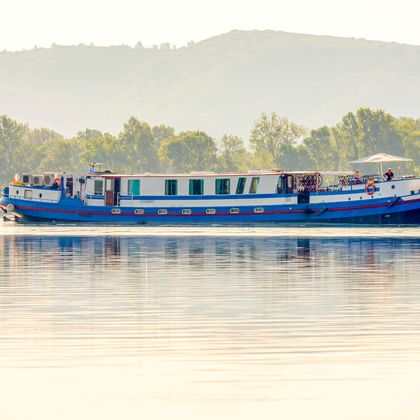 Blau-weißes Flusskreuzfahrtschiff MS Caprice fährt auf ruhigem Wasser mit Hügeln und Bäumen im Hintergrund bei sanftem Morgenlicht.
