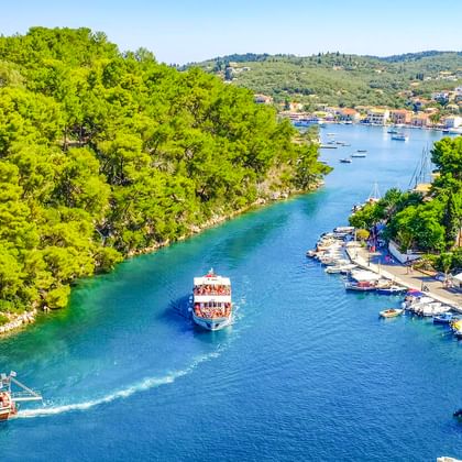 Luftaufnahme des Hafens von Gaios auf Paxos mit zwei Passagierbooten in einem schmalen Kanal, gesäumt von grünen Bäumen und Häusern.