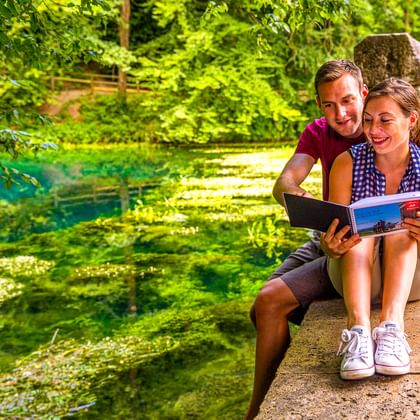 Paar sitzt am Rand des leuchtend grünen Blautopfs in Blaubeuren und liest einen Reiseführer, umgeben von üppiger Vegetation am Wasser.