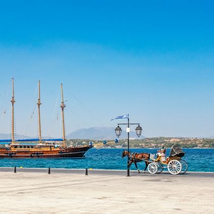 Dreimaster-Segelschiff MS Thalassa im blauen Hafenwasser. Pferdekutsche auf Uferpromenade mit Laternen und Küstenlinie im Hintergrund.