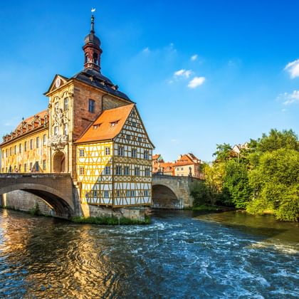 Historic half-timbered Old Town Hall in Bamberg built on a stone bridge over the Regnitz River, with tower and colorful facades under blue sky.