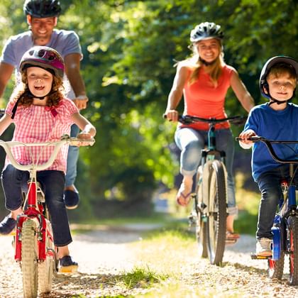 Parents and two children cycling on a tree-lined path. All wear helmets. Girl in pink shirt on red bike, boy in blue on dark bike.