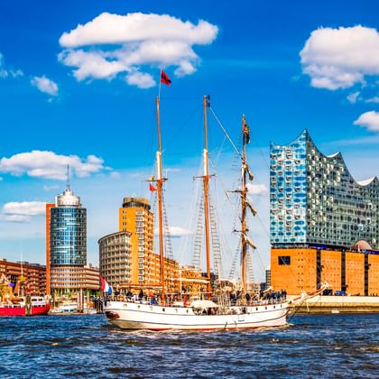 White sailing ship on the Elbe River with Hamburg's Elbphilharmonie concert hall and historic harbor buildings under a blue sky with clouds.