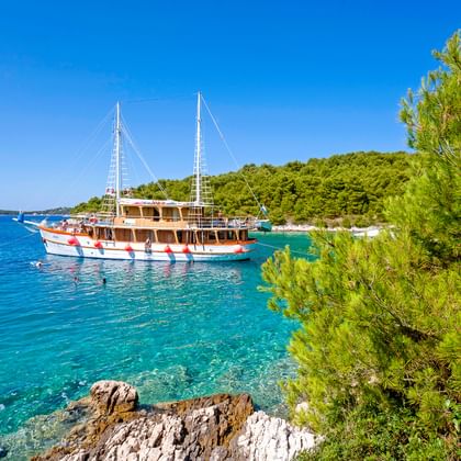 Traditionelles Holzsegelschiff MS Dalmatino vor Anker in kristallklarer türkisfarbener Bucht in Dalmatien, Kroatien, umgeben von Pinien und Felsküste.