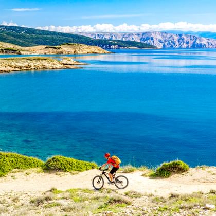 Radfahrer in roter Jacke fährt entlang Küstenweg mit Blick auf türkisfarbene Adria mit Felsklippen und Bergen in Kroatien.