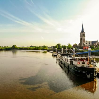 Flusskreuzfahrtschiff MS Magnifique II an einem Holzsteg vertäut. Eine Kirche mit hohem Turm steht am gegenüberliegenden Flussufer unter blauem Himmel.