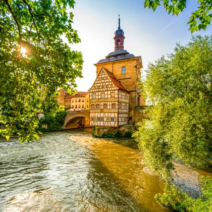 Historisches Fachwerkrathaus von Bamberg auf einer Insel in der Regnitz, umrahmt von grünen Bäumen mit durchscheinendem Sonnenlicht.